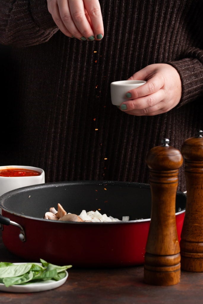 adding crushed red pepper to mushrooms and onions sauteeing in a pan