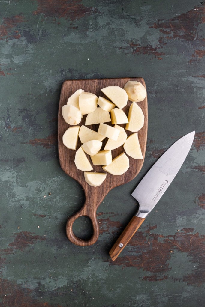 Peeled Russett potatoes that are chopped into small chunks on a chopping board with a knife laying next to it