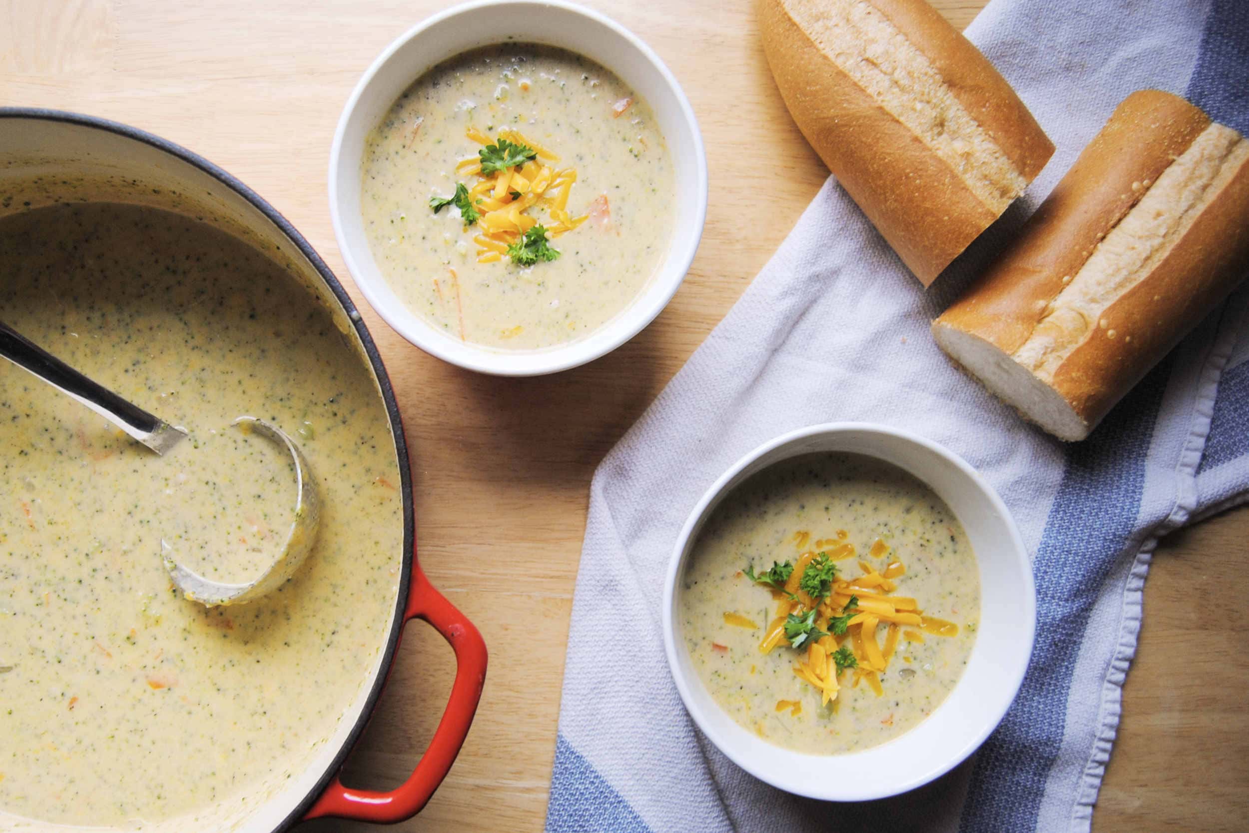 A dutch oven full of broccoli cheddar soup with a ladle and two bowls of soup with French bread