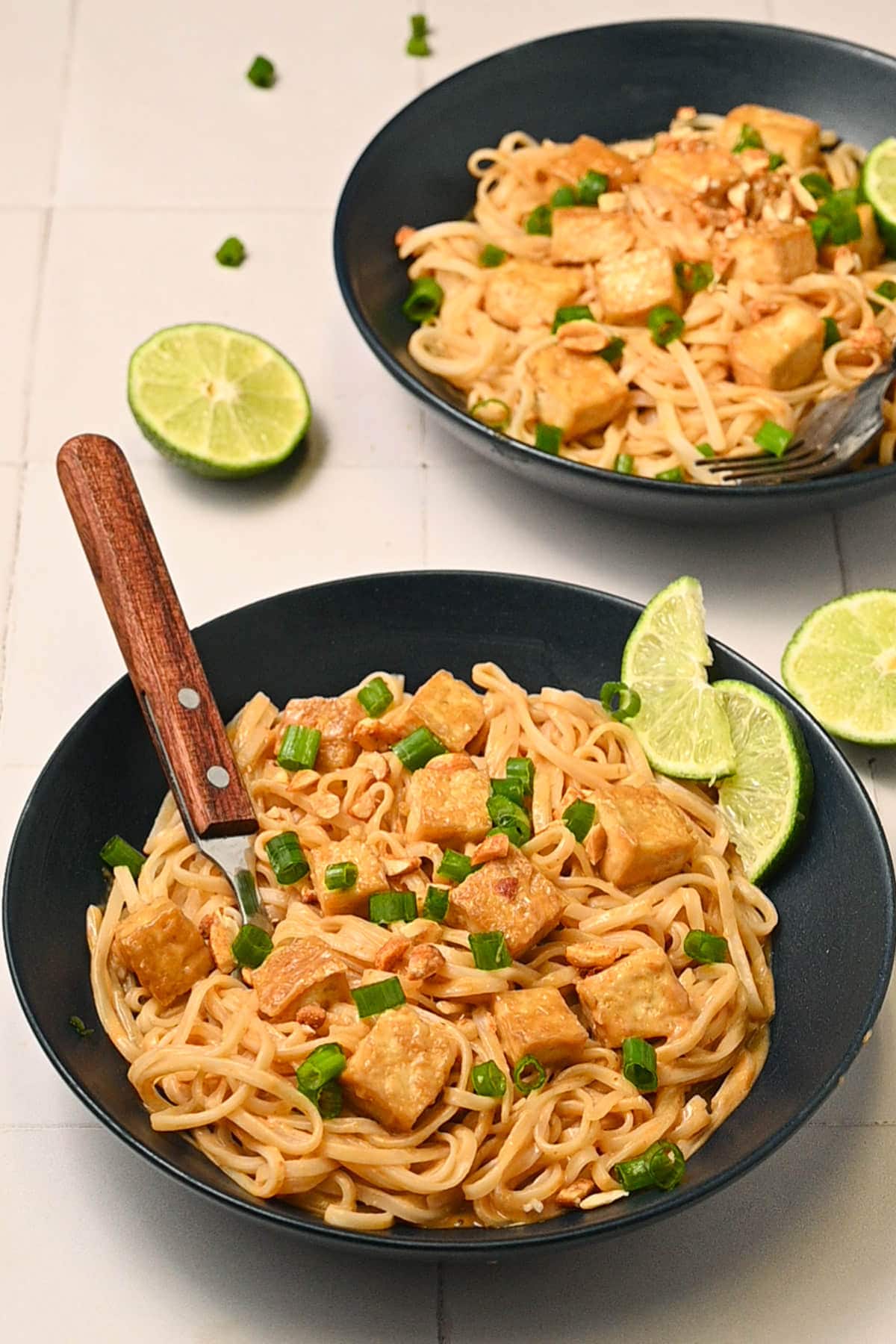 2 bowls of spicy peanut butter noodles garnished with scallions, chopped peanuts, and lime wedges.