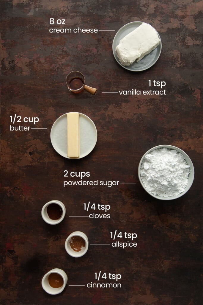 Overhead view of the ingredients needed for spiced cream cheese frosting - cream cheese, vanilla extract, butter, powdered sugar, cloves, allspice, and cinnamon.