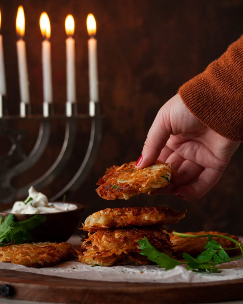 grabbing a latke from a stack with lit hanukkah menorah in the background