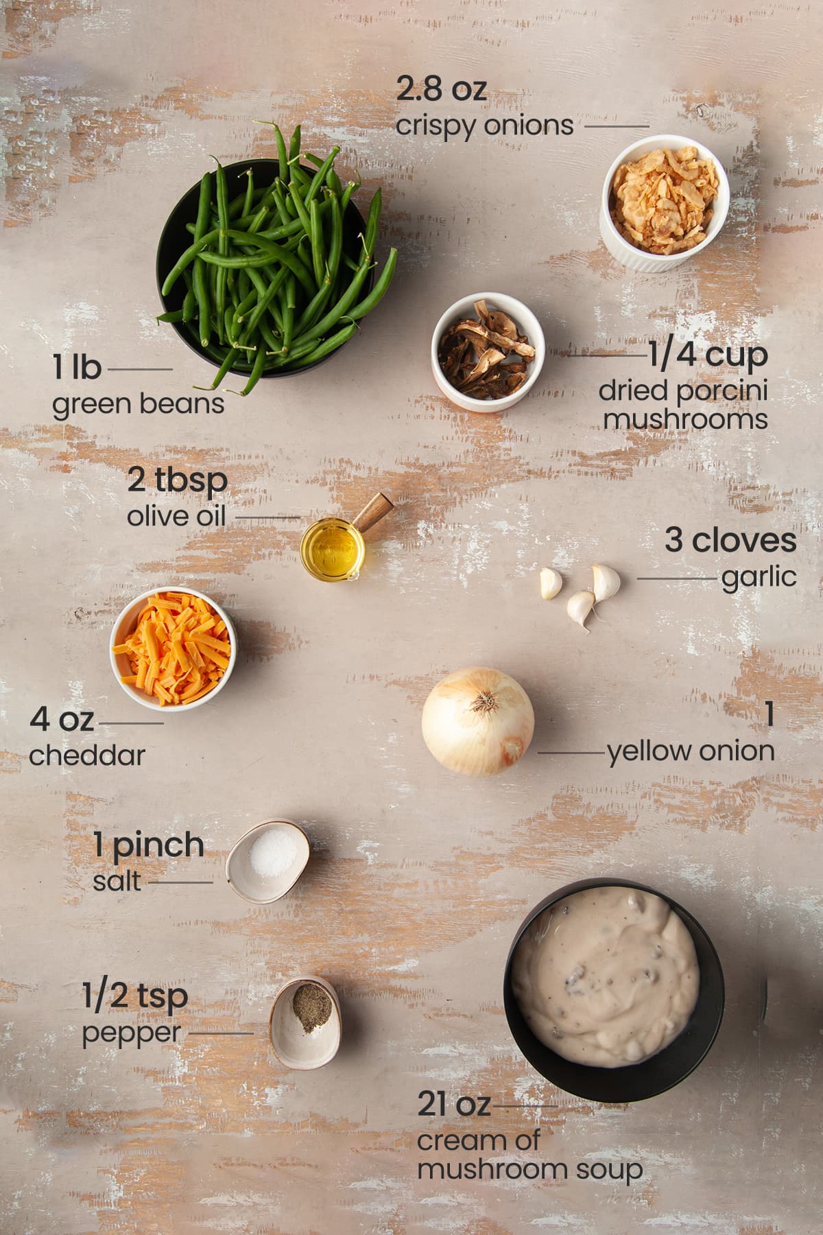 Overhead view of the ingredients needed for green bean casserole - green beans, porcini mushrooms, cheddar cheese, olive oil, garlic, onion, salt, cream of mushroom soup, and black pepper.