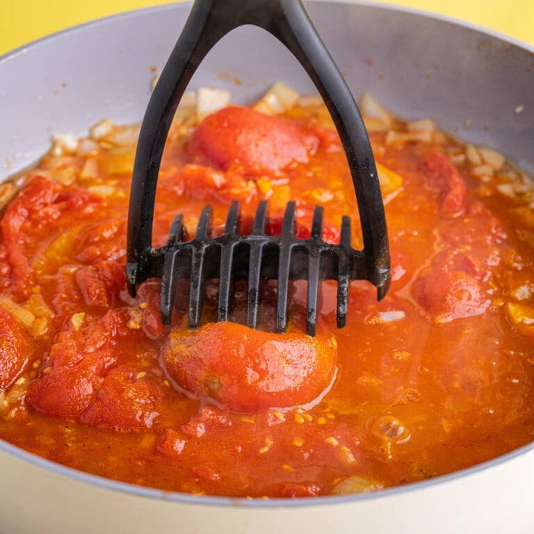 Using potato masher to crush whole peeled plum tomatoes in a pan