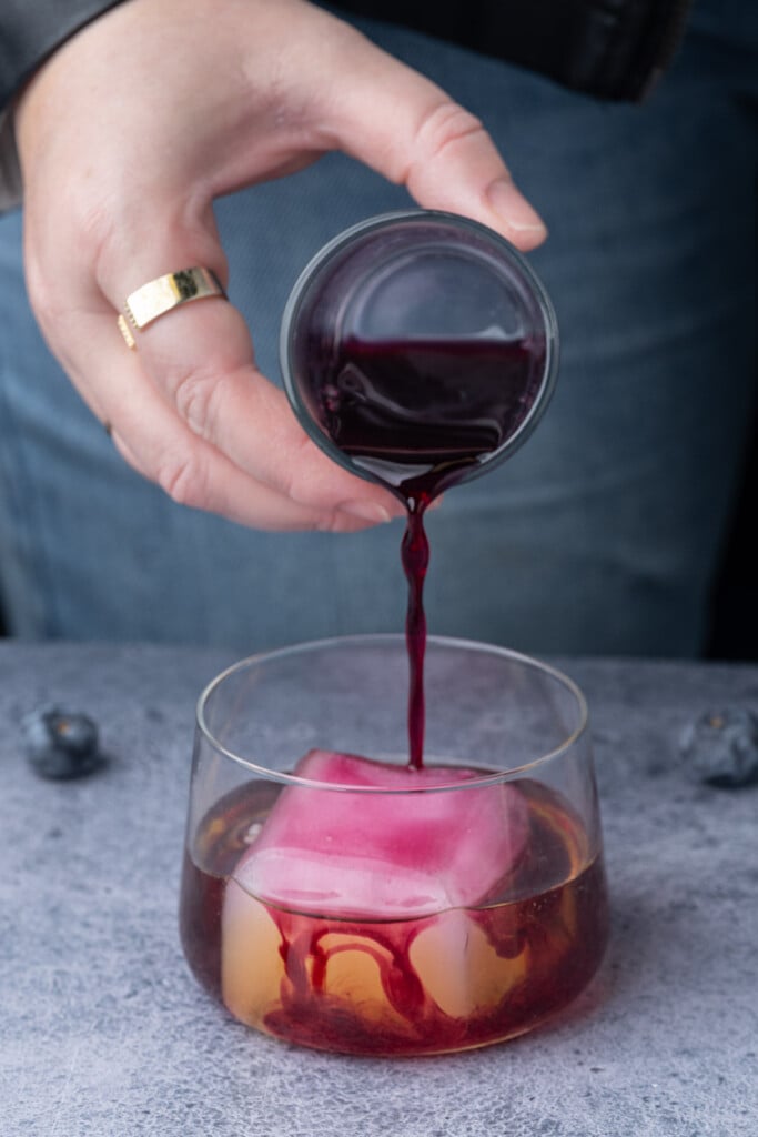 Pouring blueberry simple syrup into a glass with whiskey and a big block of ice. 
