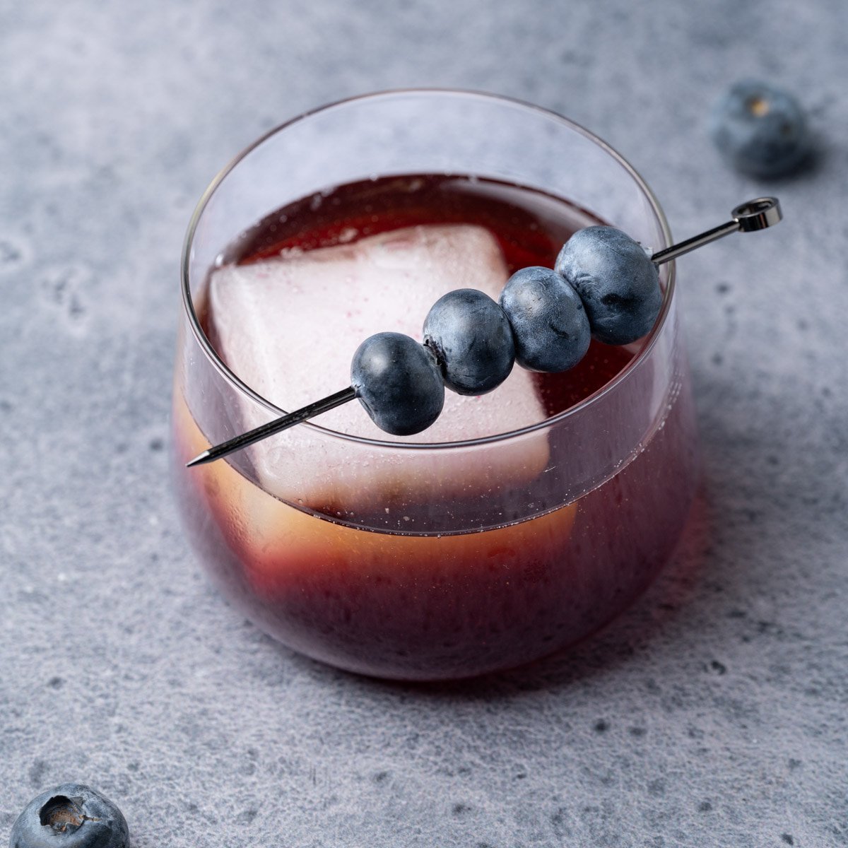 Lowball glass with a vibrant blue whiskey cocktail garnished with fresh blueberries.