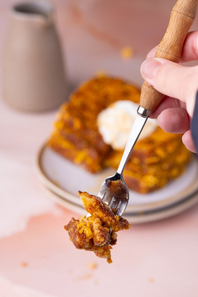 Holding up a forkful of crispy Cap'n Crunch French Toast to the camera with a full stack topped with whipped cream in the background. 