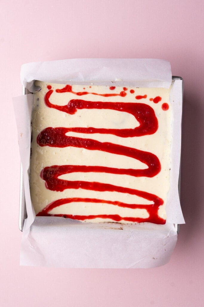 Pouring raspberry sauce onto cheesecake brownies in a zig zag. 