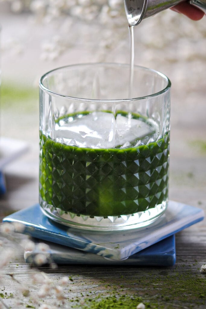 Adding vanilla liqueur to matcha vodka in a small glass.
