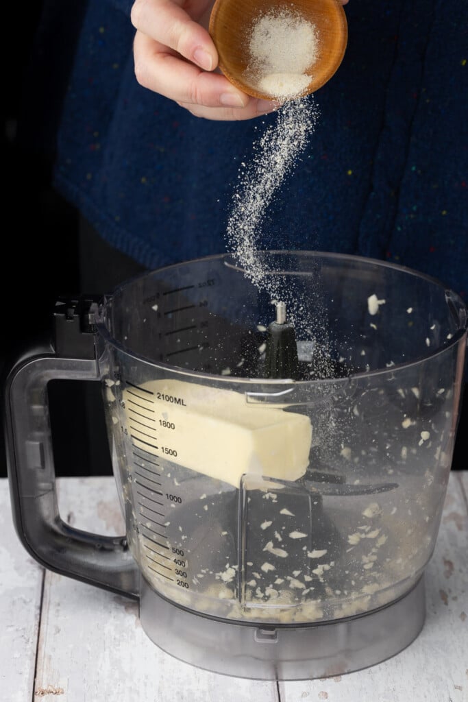 Adding garlic salt to a food processor with softened unsalted butter and fresh minced garlic.