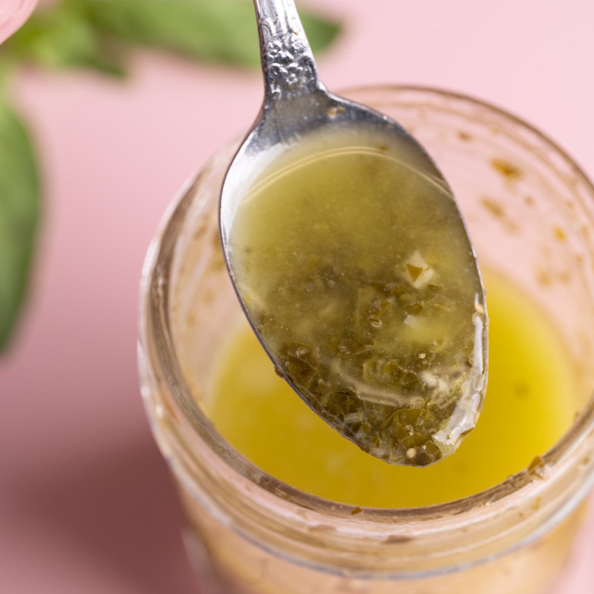 Close up of a spoon with homemade pesto vinaigrette dripping off of it into a glass jar.