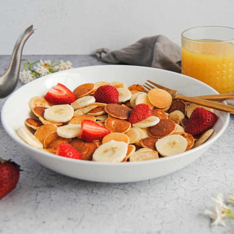 Baby Banana Pancakes in a serving bowl with utensils, juice, and syrup bottle