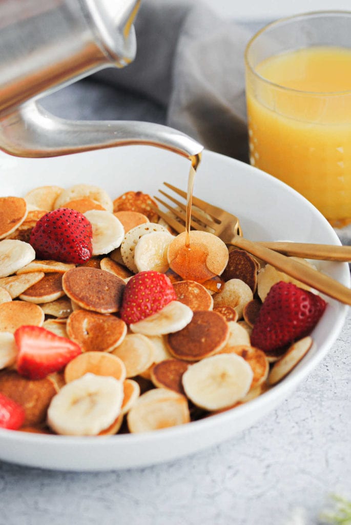 pouring syrup onto a bowl of baby pancakes with strawberries