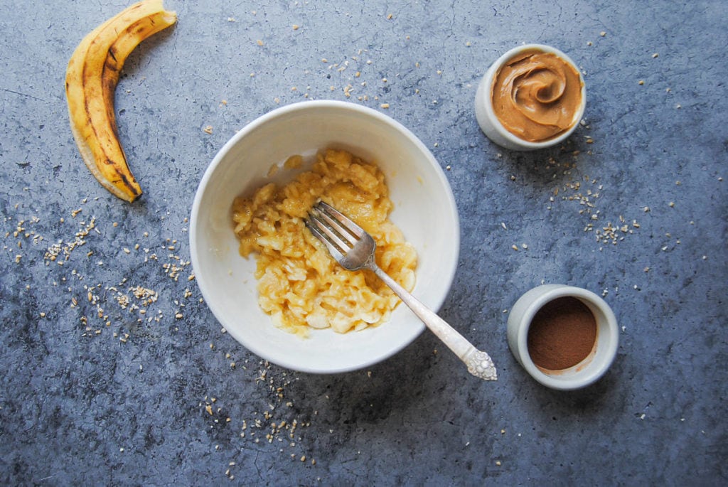 bowl with banana in it being mashed by a fork surrounded by cinnamon and peanut butter
