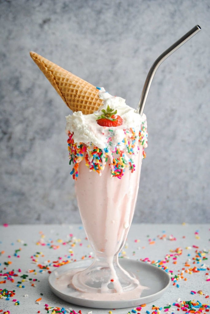 strawberry milkshake surrounded by rainbow sprinkles garnished with strawberry and a steel straw