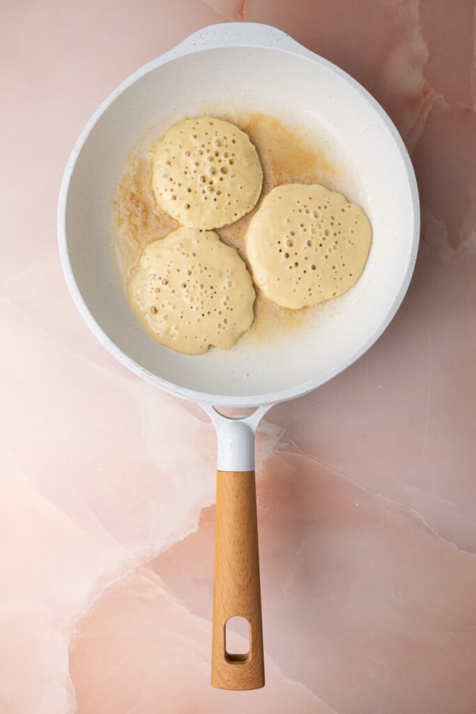 Frying three pancakes in a pan with hot butter, showing that they are ready to flip because there are bubbles in the batter. 