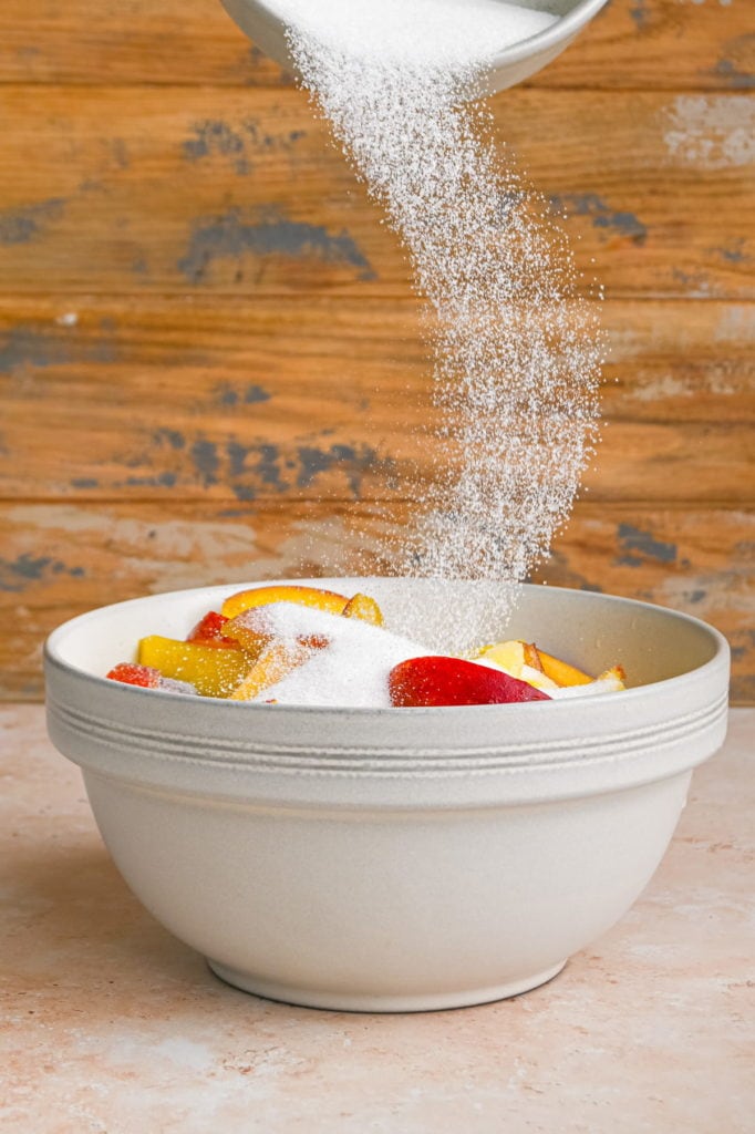 adding sugar to mixing bowl filled with sliced peaches and mangoes