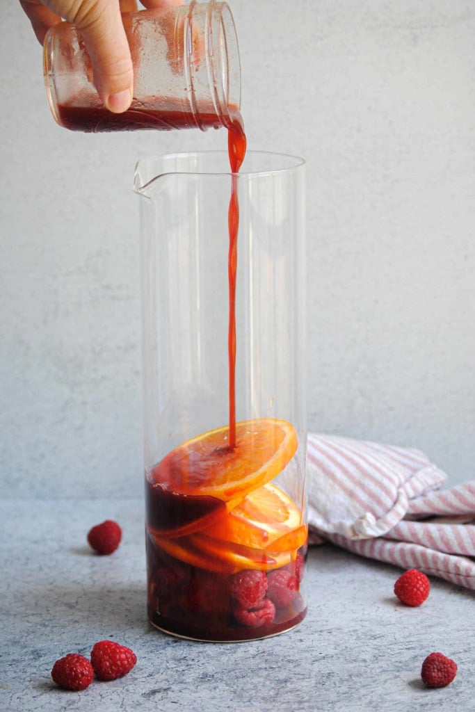pouring raspberry simple syrup into a pitcher filled with sliced oranges and more raspberries