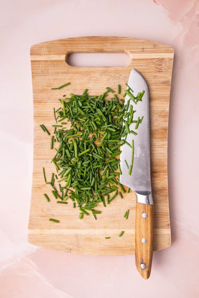 Diced chives on a wooden cutting board with a sharp knife laying on top. 