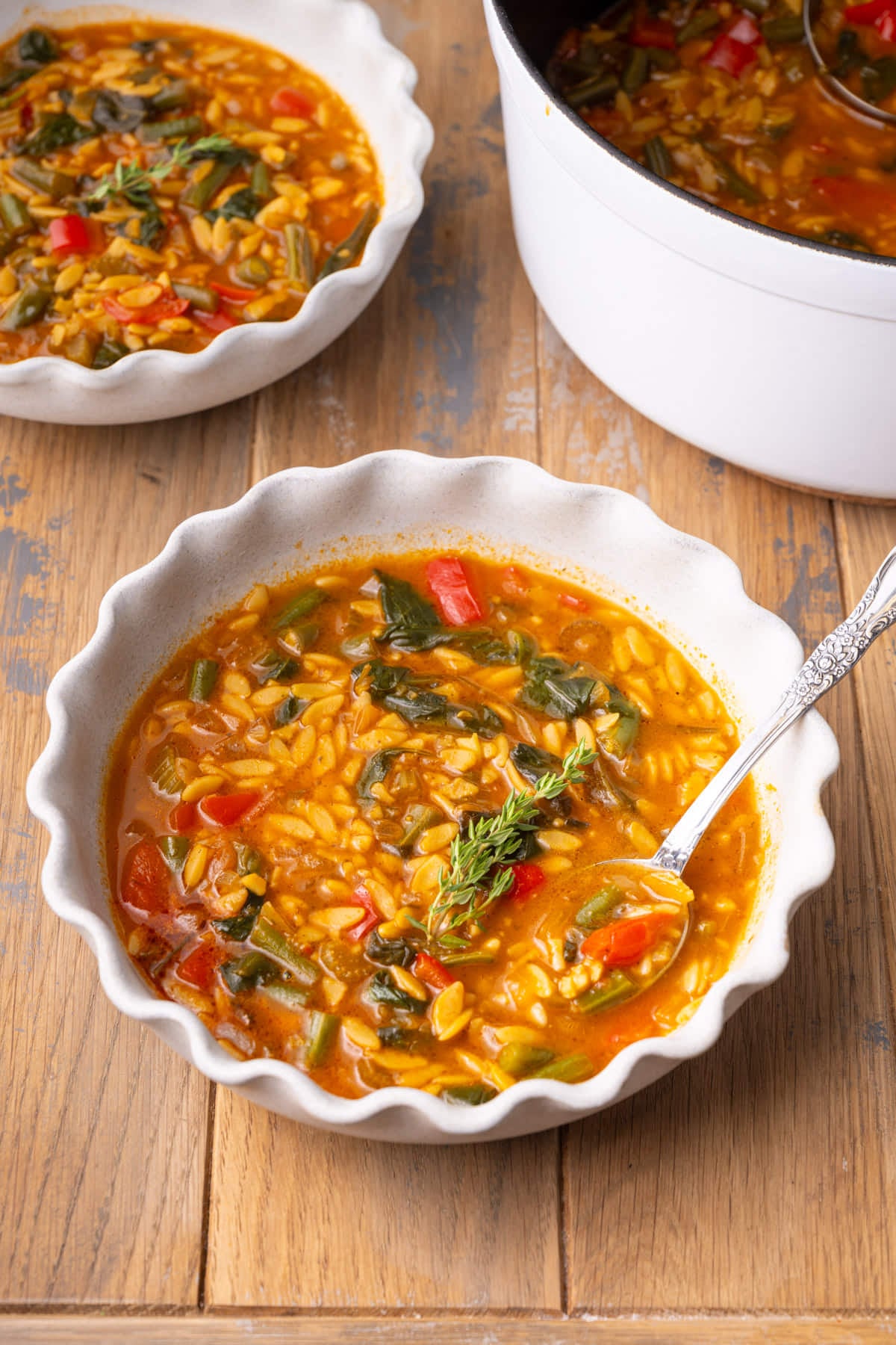 Bowl with scalloped edges filled with a hot soup with vegetables and orzo, with another bowl and the pot in the background.