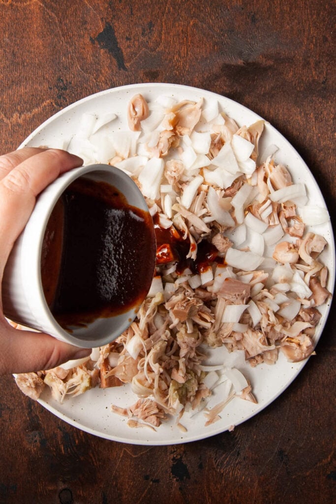 Adding BBQ sauce to a plate of pulled BBQ jackfruit and diced onions.