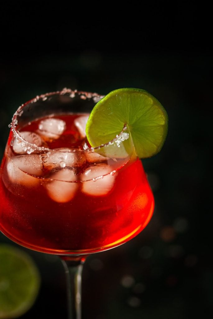 Close Up of Salt Rimmed Glass With Ice and Red Cocktail Garnished With Lime