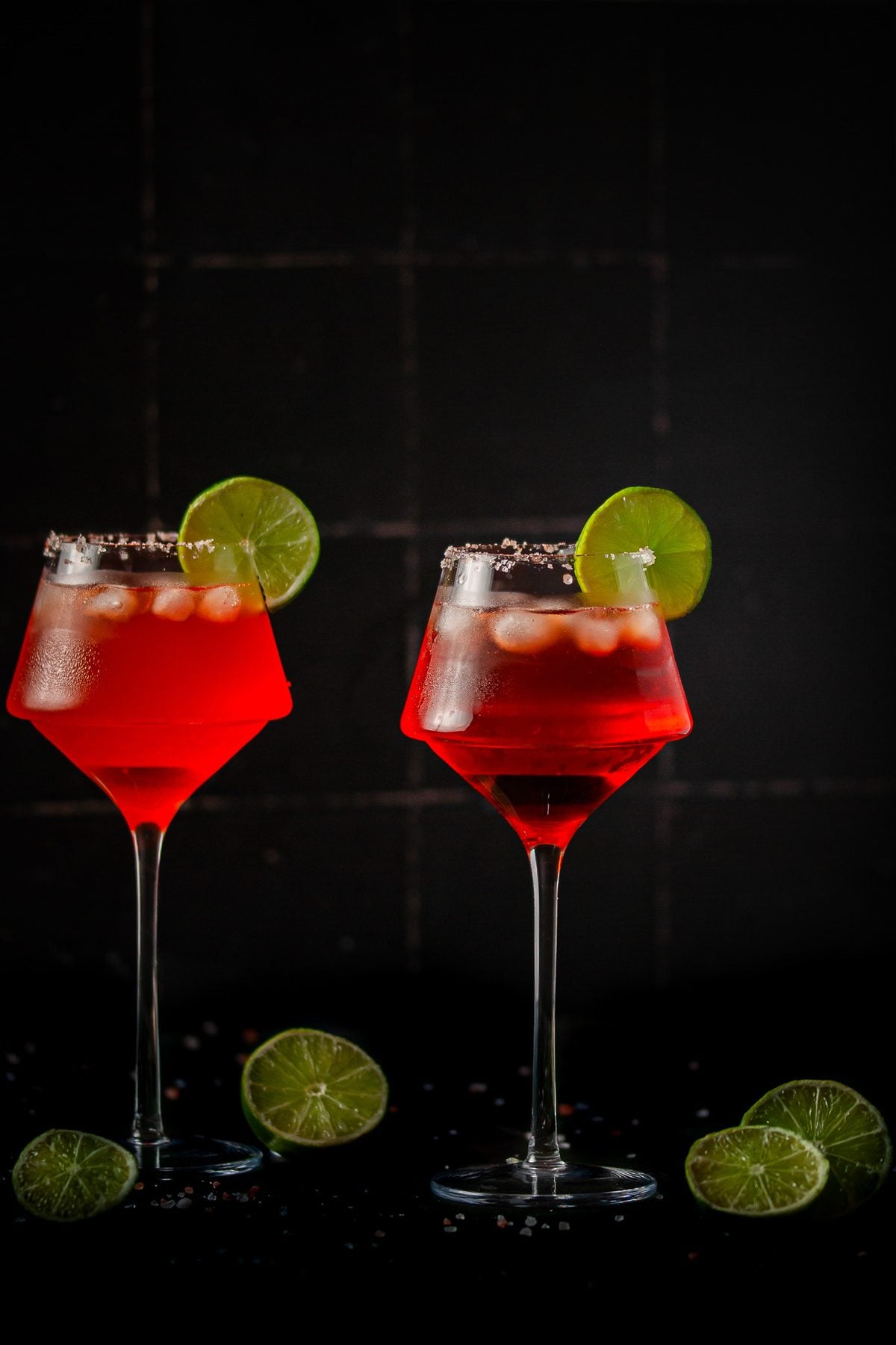 Moody picture of two bright red margaritas against a black tile background.
