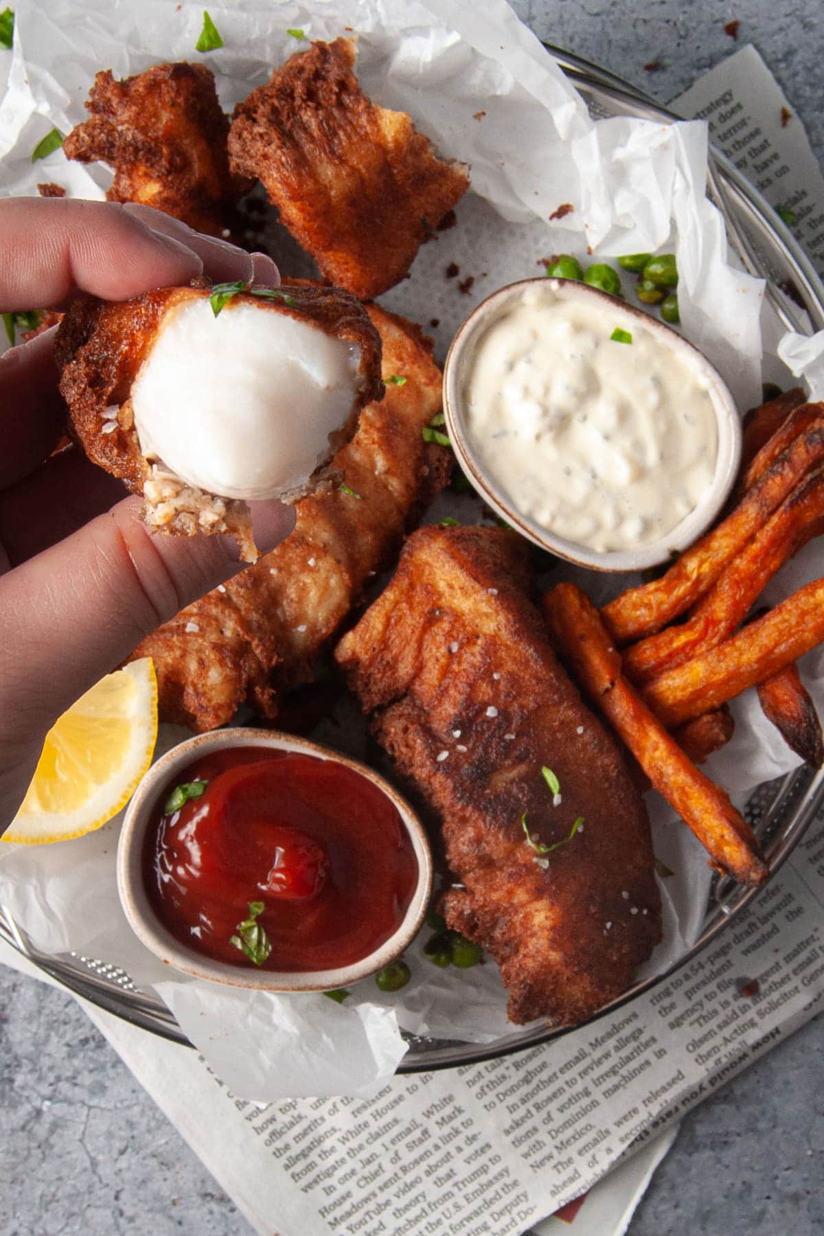 holding piece of beer battered fish close to the camera with platter of fish in the background