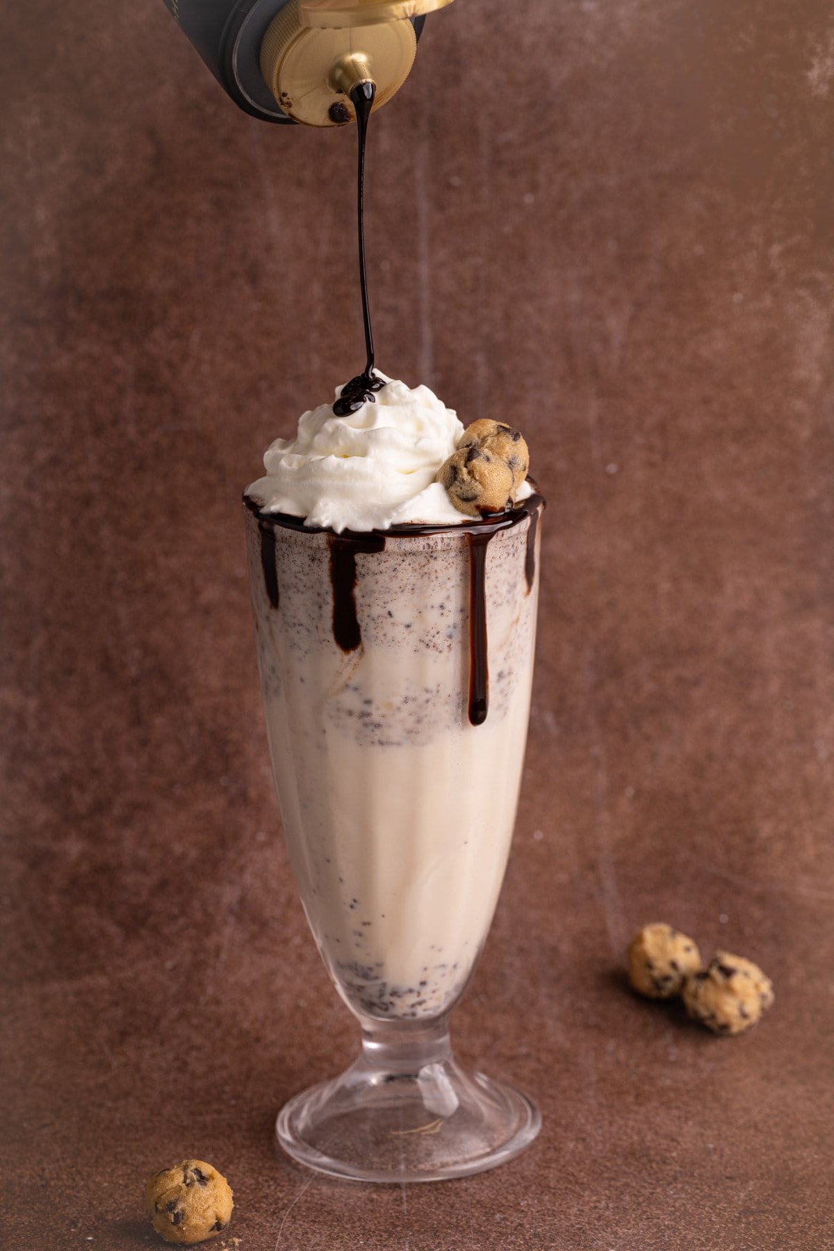 Drizzling chocolate sauce on top of milkshake topped with whipped cream and cookie dough. 