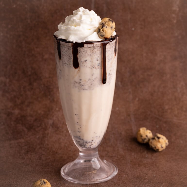 Cookie dough milkshake in an old fashioned fountain glass topped with whipped cream, two cookie dough balls, and finished with a chocolate sauce rim.