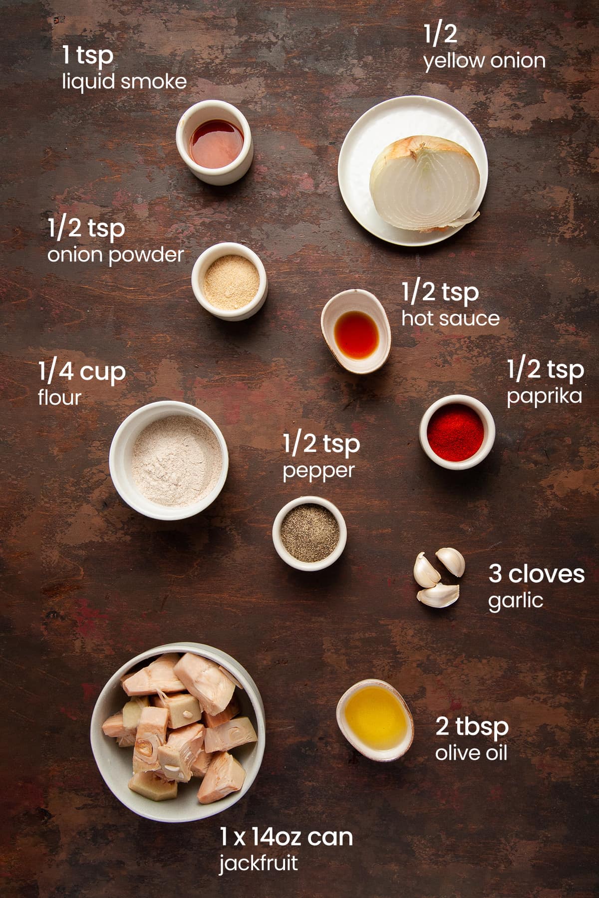 Overhead view of the ingredients needed for oven baked jackfrui burgers - hot sauce, 1/2 an onion, onion powder, liquid smoke, paprika, flour, pepper, garlic, jackfruit, and olive oil.