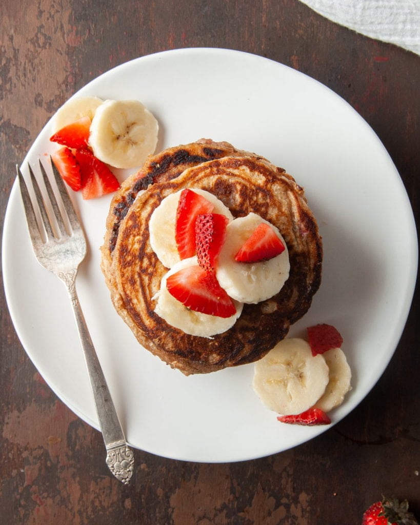 overhead shot of stack of pancakes on a plate with fork and sliced bananas and strawberries