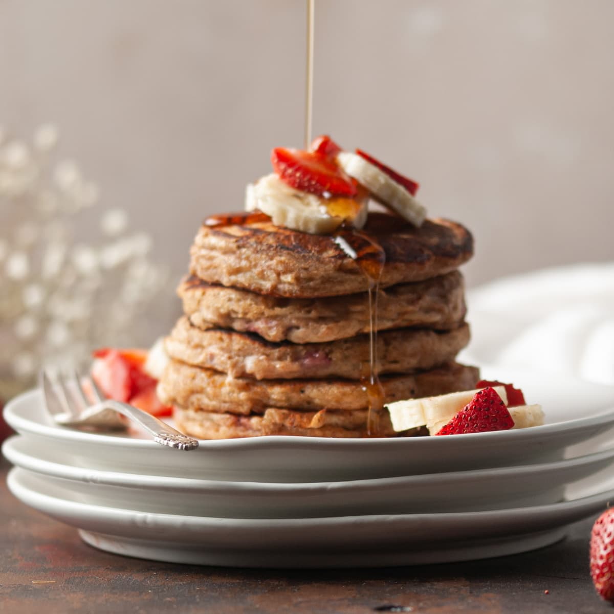 stack of whole wheat pancakes with bananas and strawberries and syrup being drizzled on top