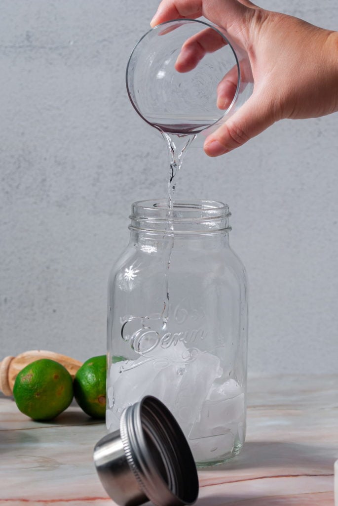 pouring vodka from a small glass container into a big mason jar with ice