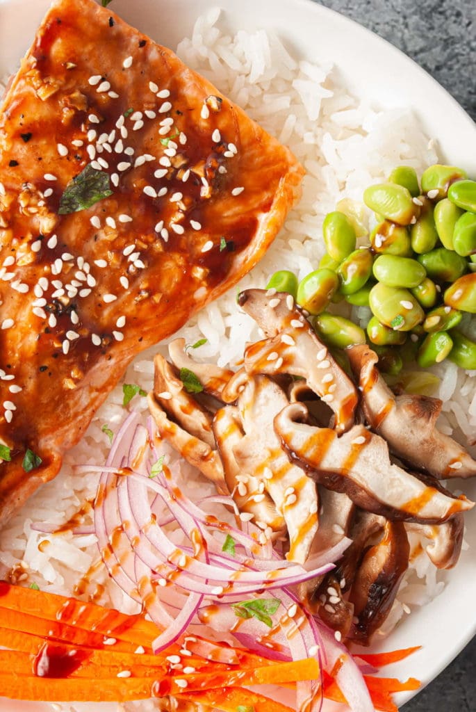 close up of rice bowl with teriyaki glazed salmon, shiitake mushrooms, edamame, red onion, carrots. and teriyaki glaze on top