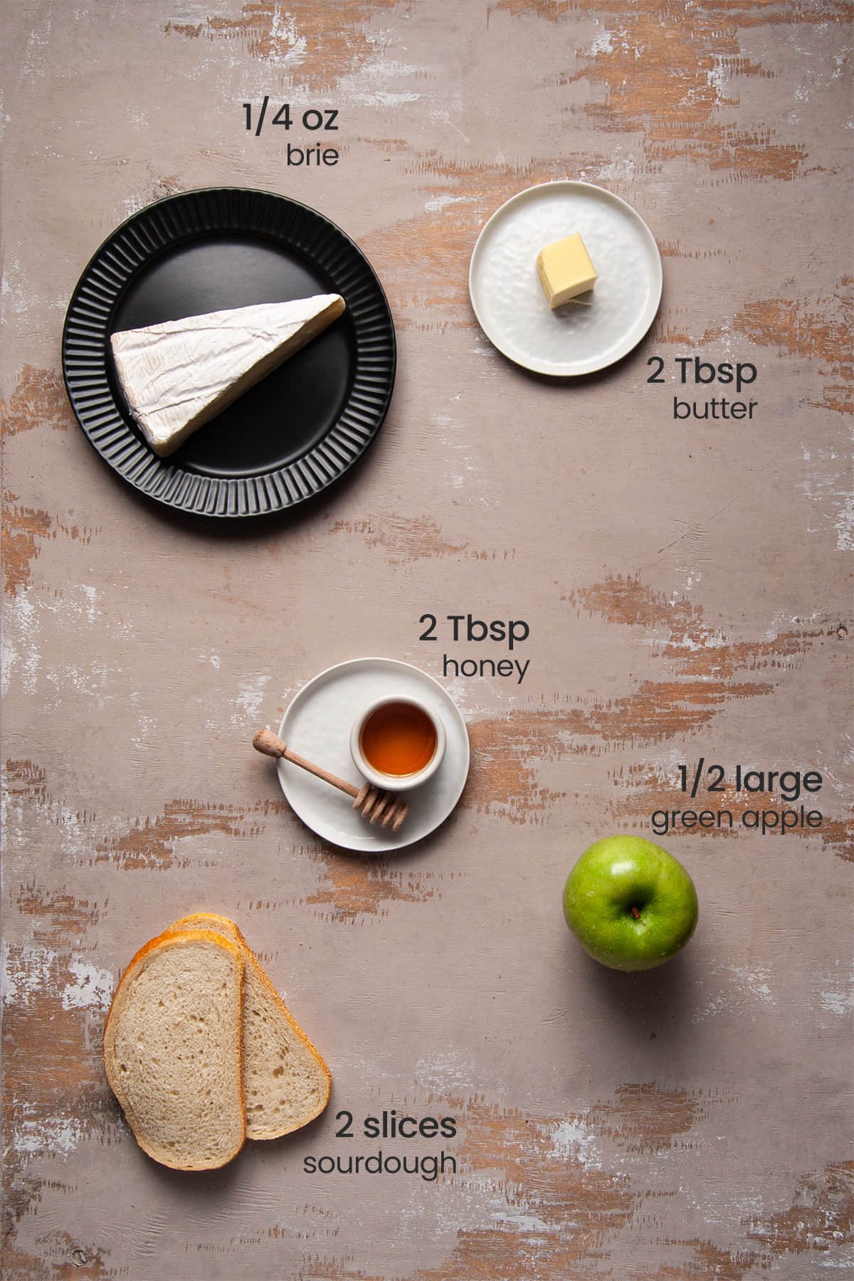 Overhead image of all of the ingredients needed for Brie Apple Grilled Cheese labeled with their amounts, including brie cheese, salted butter, honey, a green apple, and sourdough bread.
