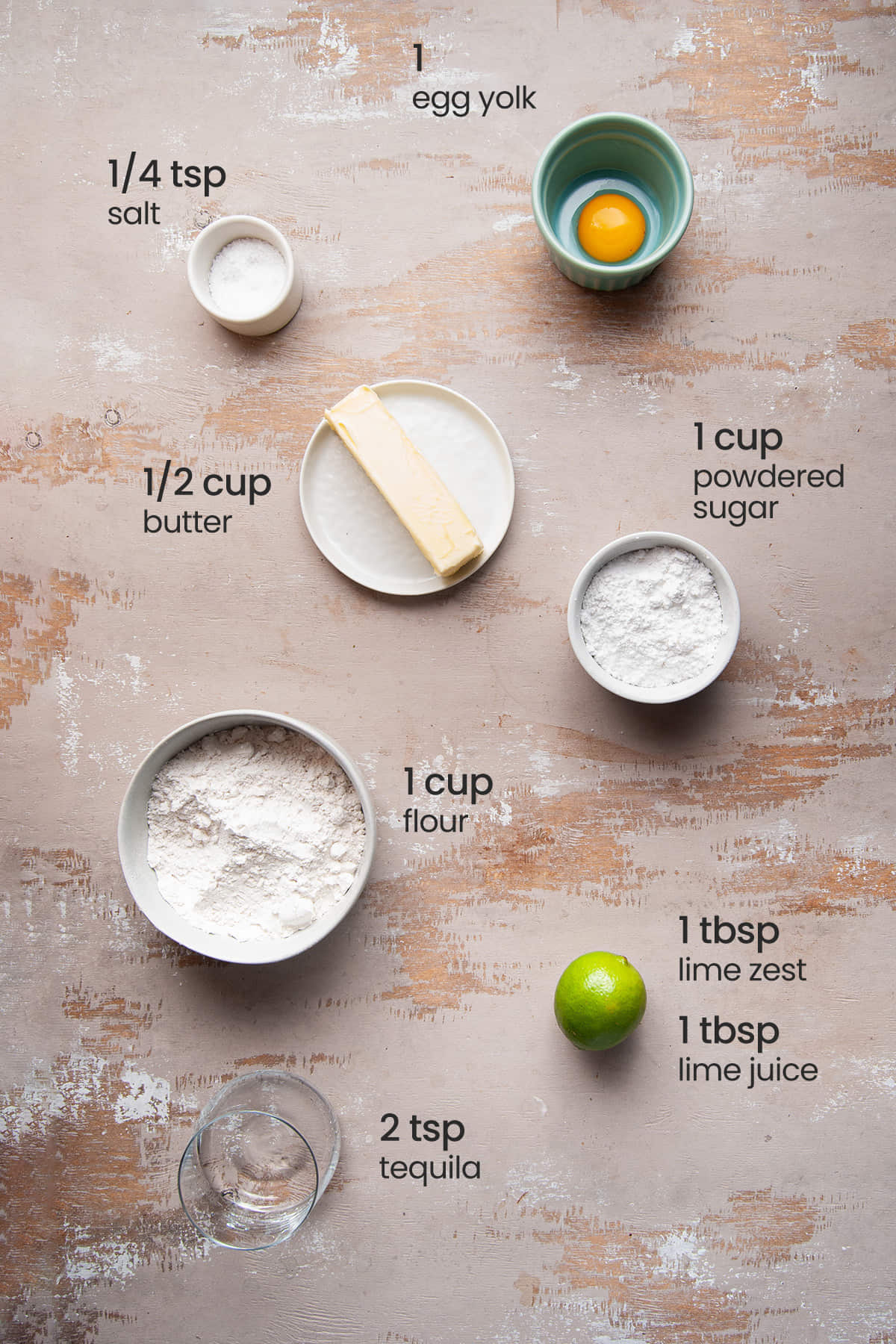 Overhead view of all ingredients needed for Margarita Cookies with Tequila Lime Glaze - egg yolk, salt, butter, powdered sugar, flour, lime, and tequila.