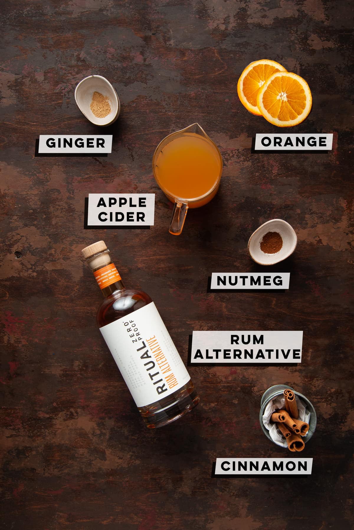 Overhead picture of the ingredients in an apple cider spiced rum mocktail - ginger, apple cider, orange, nutmeg, rum alternative and cinnamon