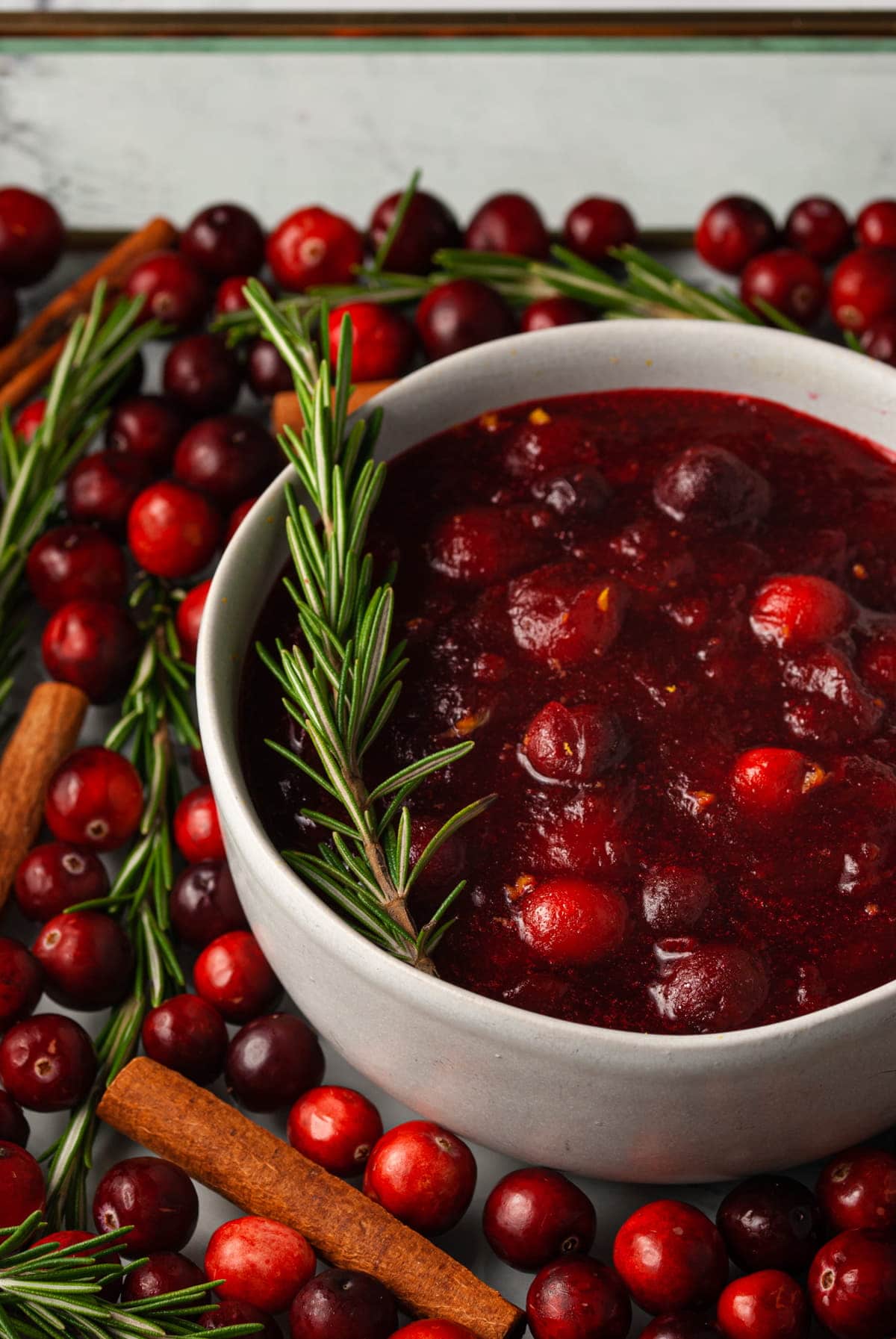 bowl of cranberry sauce with sprig of rosemary garnish, surrounded by fresh cranberries