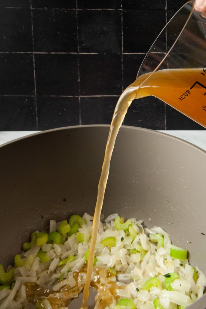 adding veggie broth to a large pot with cooked onions, celery, and fresh herbs