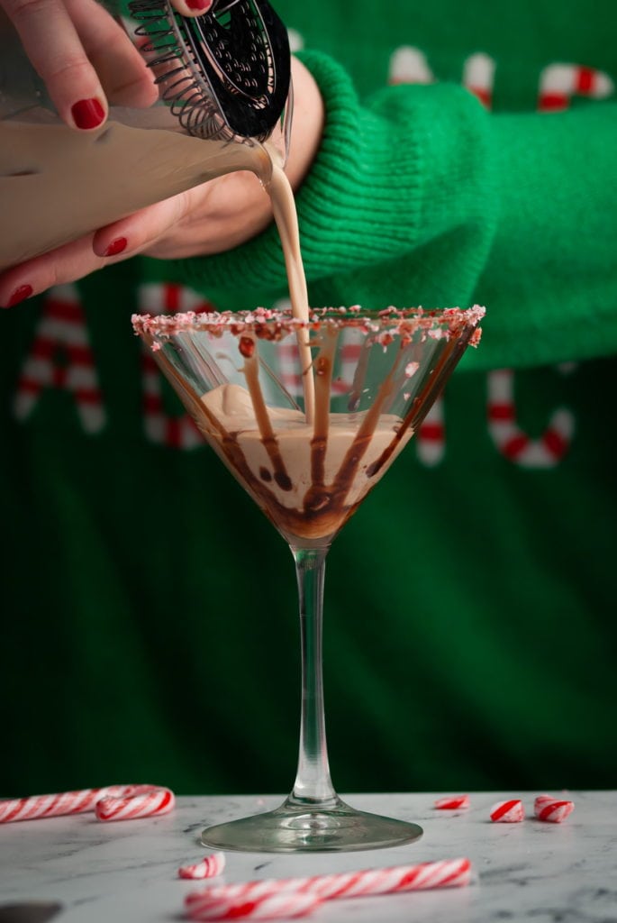 pouring peppermint mocha martini into a martini glass garnished with candy cane and chocolate syrup