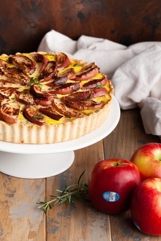 a sweet and savory quiche on a cake stand surrounded by fresh apples