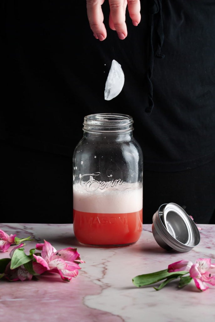 adding ice to a cocktail shaker with a pink cocktail and foam