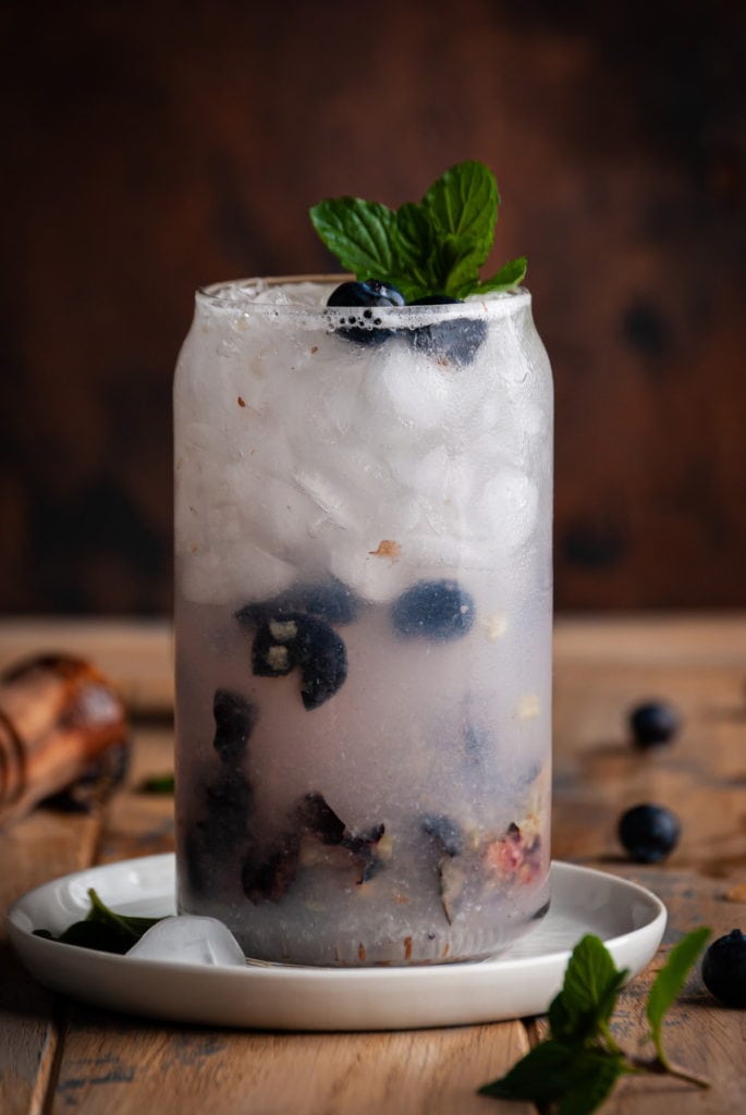 straight on shot of glass filled with a blueberry gin cocktail with ice and mint garnish
