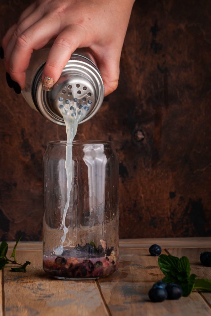 pouring gin cocktail into a glass with muddled blueberries and mint