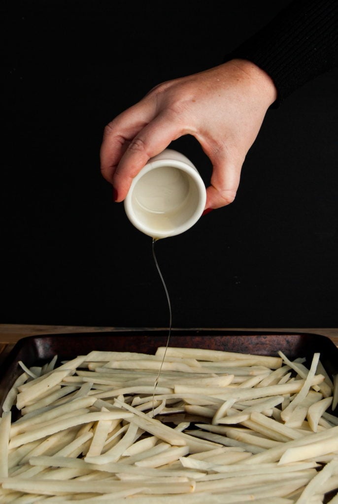 adding olive oil to raw fries on a baking sheet