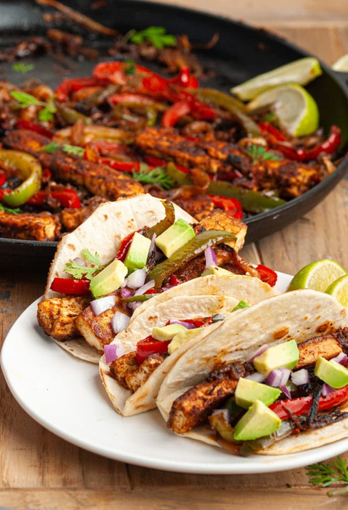 3 tofu fajitas assembled on a plate with pan of extra fajita ingredients behind it