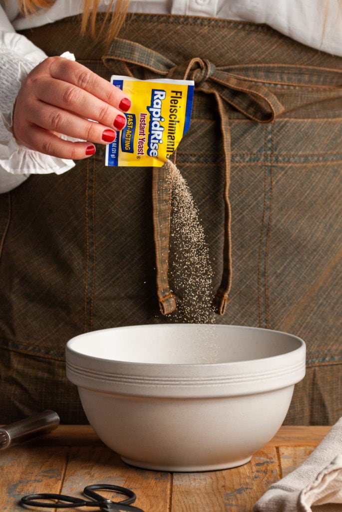 Adding a packet of instant yeast to a large mixing bowl with hot water and brown sugar