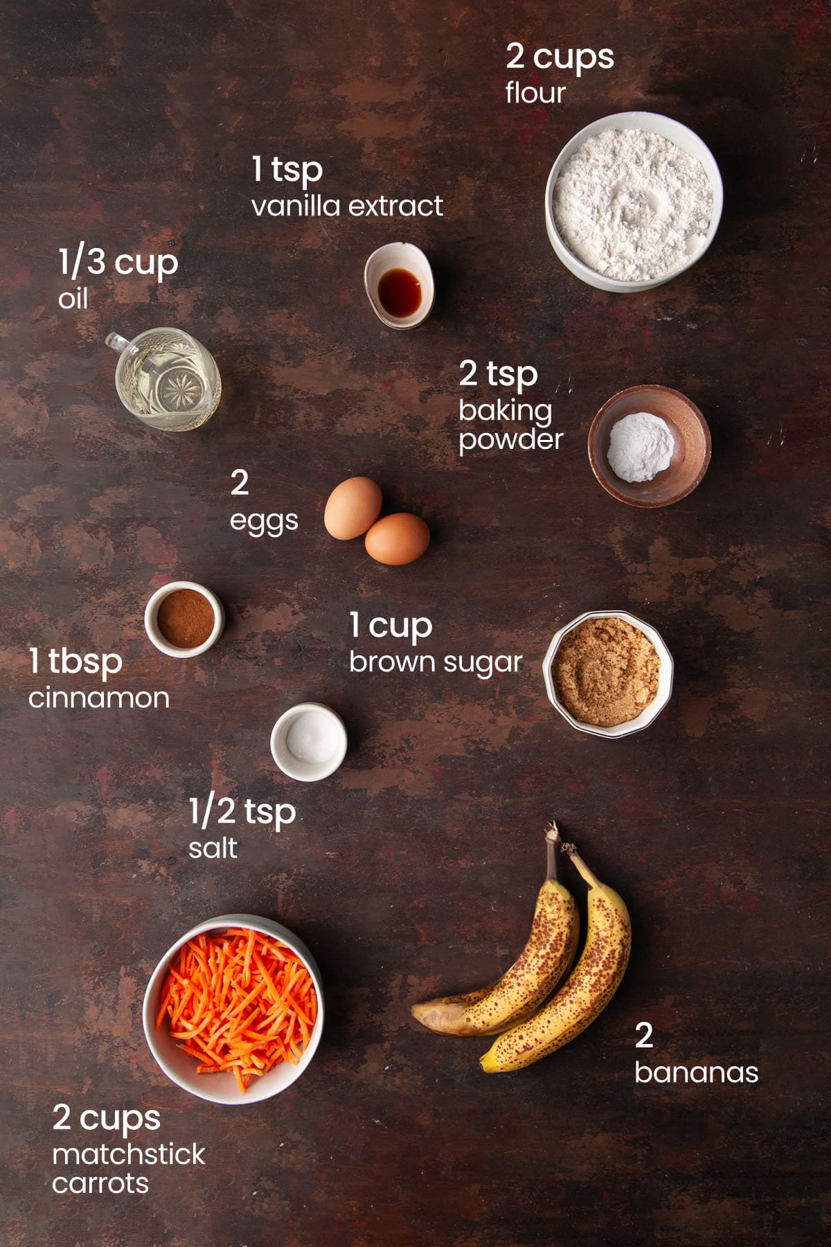Overhead view of the ingredients needed for banana carrot muffins - flour, vanilla extract, vegetable oil, baking powder, eggs, cinnamon, salt, brown sugar, carrots, and bananas.