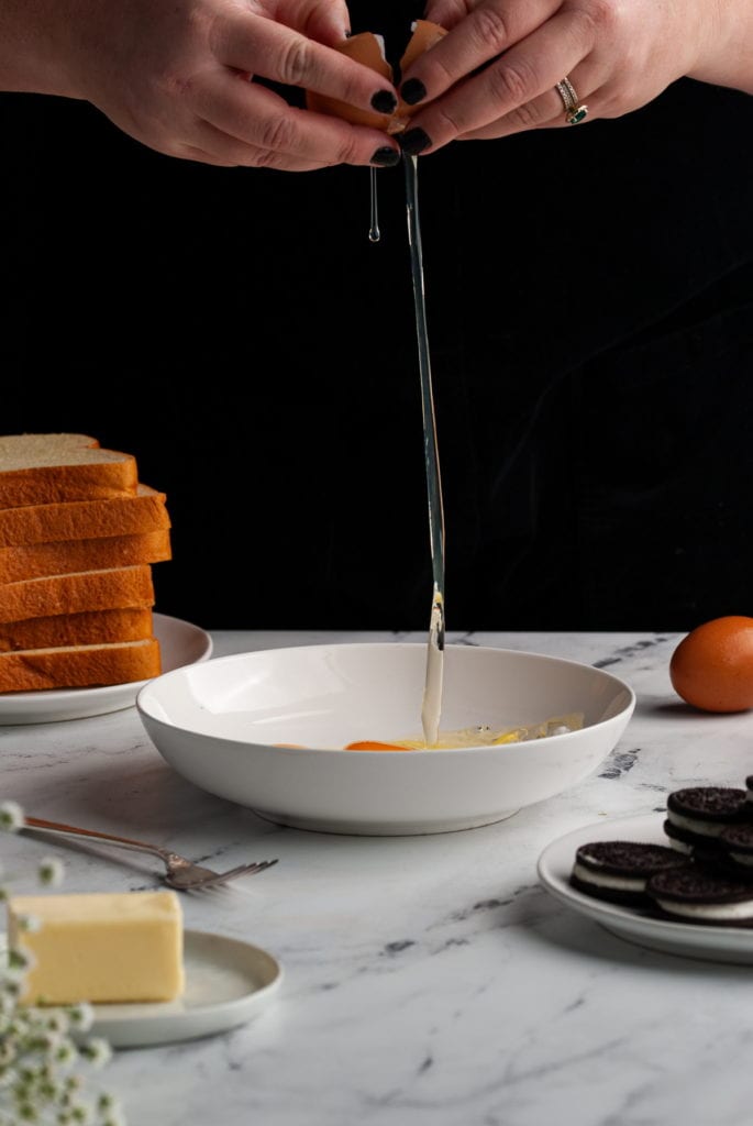 cracking an egg into a shallow bowl surrounded by ingredients for oreo french toast